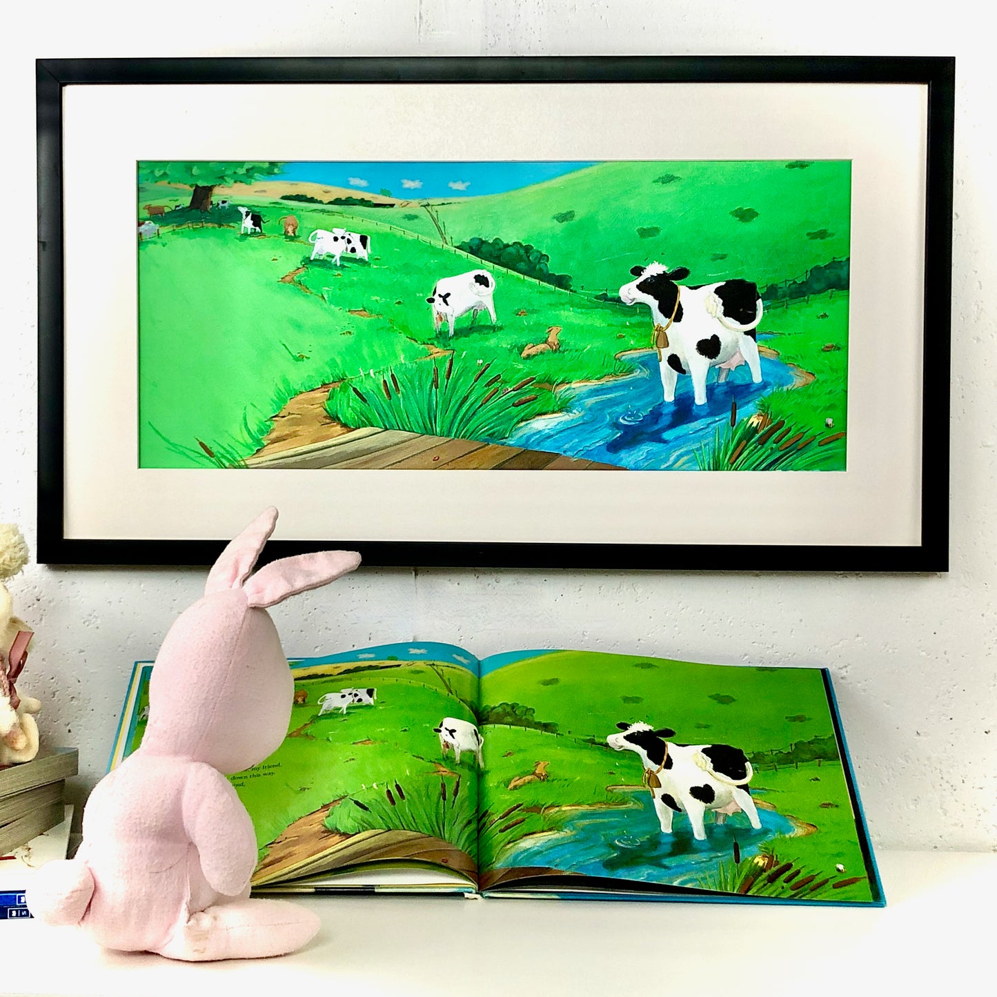 A Bunny sits in front of a book, reading the story, Buttercup's Lovely Day, which features a loving cow.  On the wall behind her is a framed cow picture from one of the pages of the books. 